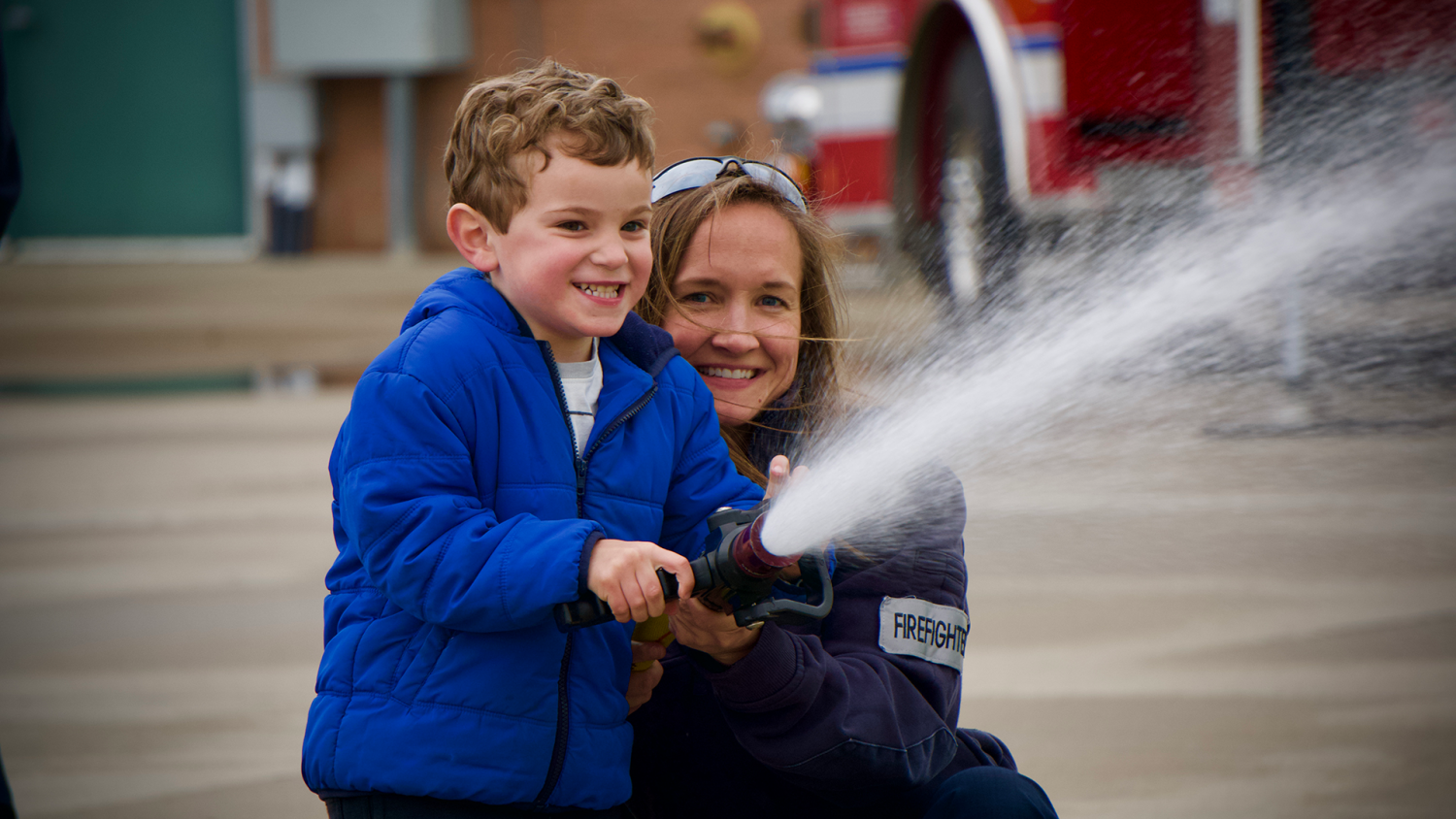 Firefighter assists young boy with holding the fire hose as it sprays.