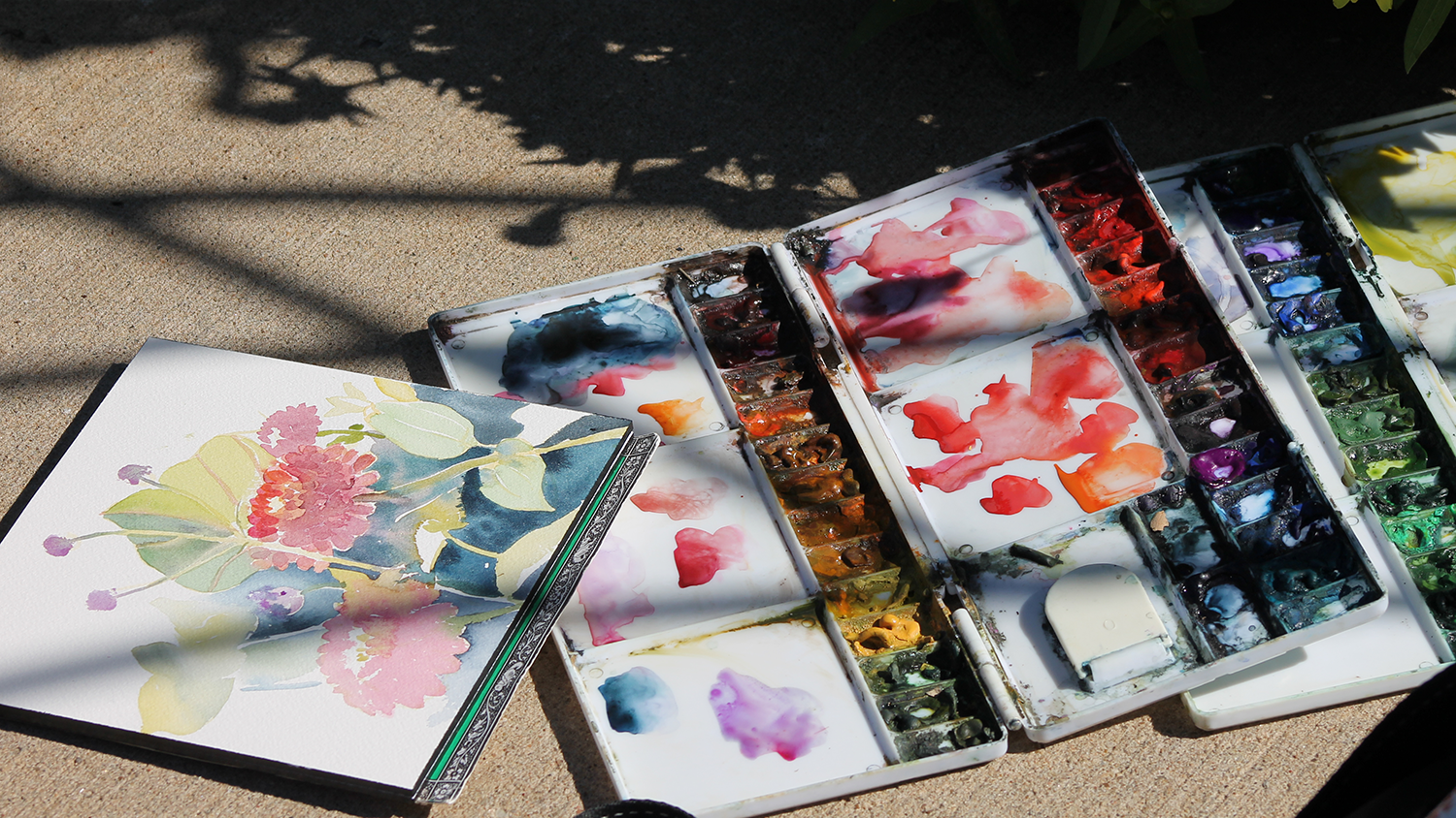 A painting of colorful flowers in a sketchbook lays on the sidewalk next to an open container of watercolors paints.