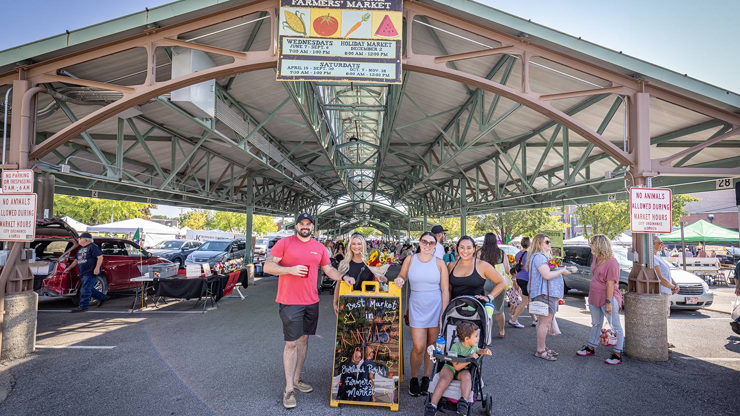 A family poses in front of the Farmers' Market Pavilion on a sunny day.